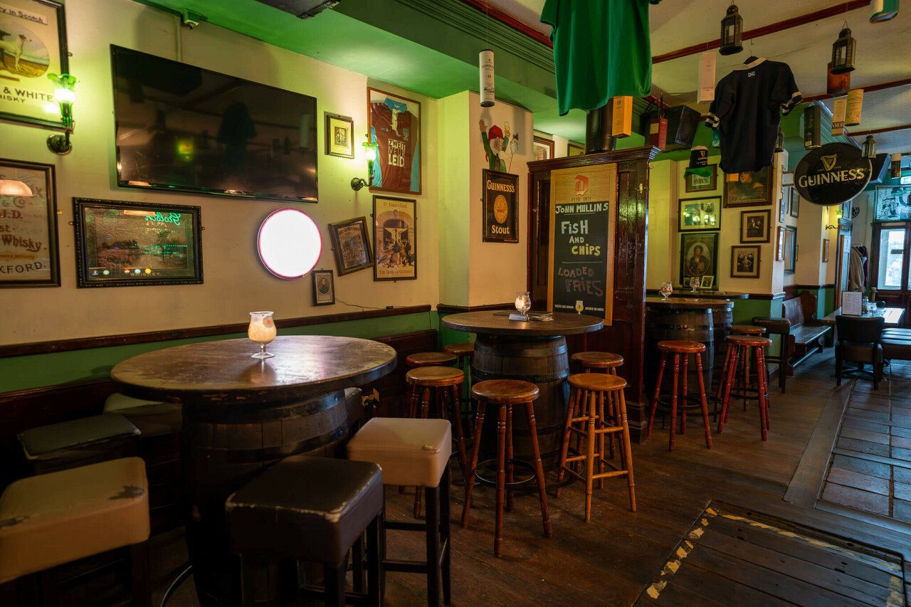 _dsc1930-enhanced-nr.jpg A cozy pub interior features wooden barrel tables surrounded by tall stools, with framed art and signage adorning the walls. A television is mounted on one wall, and a chalkboard menu highlights fish and chips. Ambient green lighting adds a warm atmosphere.